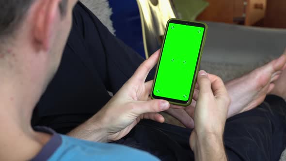 A Man on the Couch With a Phone in His Hands With a Green Screen Back View