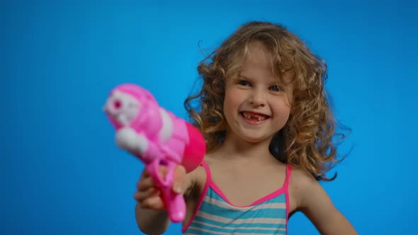 Little Blond Curly Girl with Pink Water Gun Smiles in Studio on Blue Background