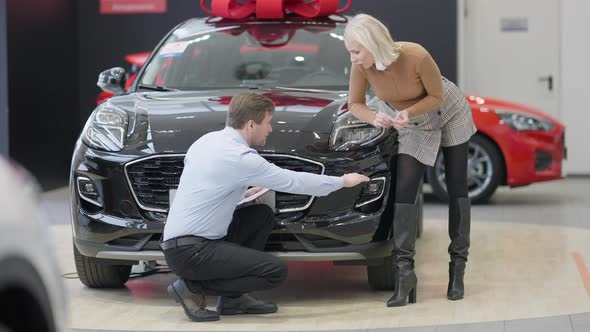 Wide Shot of Car Dealer Sitting on Hunkers Talking with Gorgeous Blond Woman Choosing Vehicle in