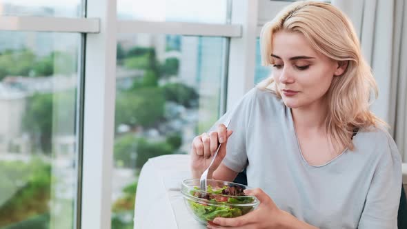 Young blonde smiling woman sitting near the panoramic window and eating healthy salad