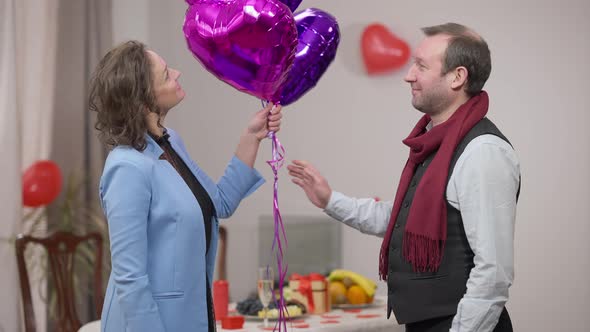 Side View of Loving Adult Man Passing Heart Shape Balloons to Shy Smiling Woman Indoors on Valentine