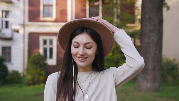 Smiling Modern Woman in Stylish Hat Walking Nearby Beautiful Rich Houses and Listening Music