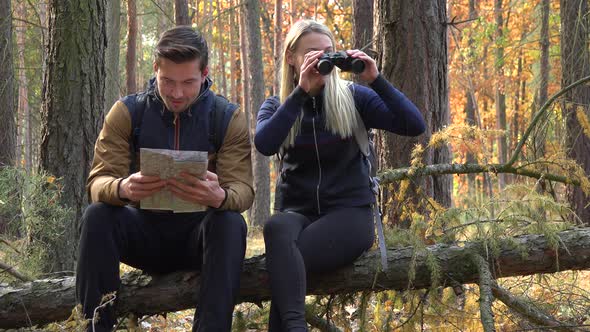 A Hiking Couple Sits on a Broken Tree in a Forest, Man Reads Off a Map and the Woman Looks Around