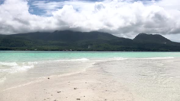 Clear blue ocean waves washing over a white sand bar with a lush green tropical island with white cl