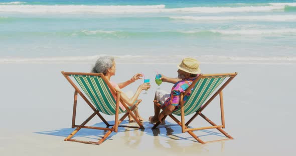 Rear view of active senior African American couple toasting drinks on deckchair at beach 4k