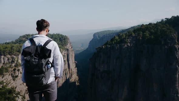 A Traveler Guy with a Backpack Stands on a Cliff Overlooking the Canyon