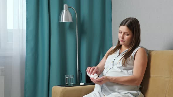 Pregnant Woman Drinks Medication with Water While Sitting on the Couch