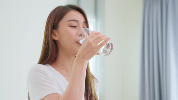 Asian attractive woman sitting on bed, drink a glass of water after wake up in bedroom.