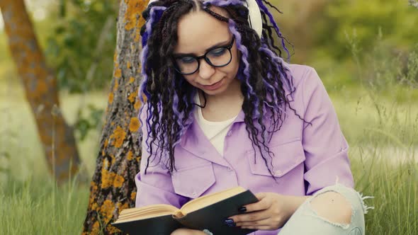 Young Woman in Glasses Headphones Reading Book Sitting Near Tree in Summertime