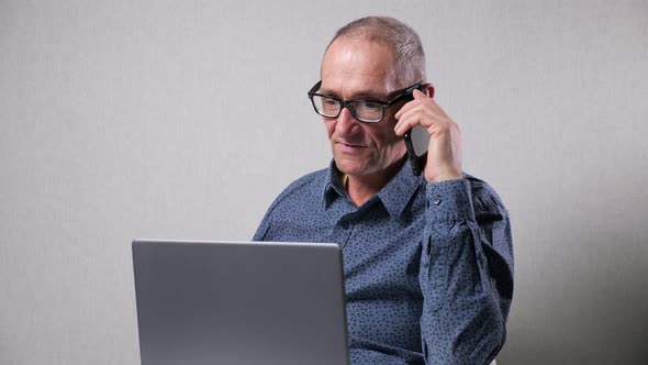 Elderly Man Talks on Mobile Phone Working on Laptop at Table