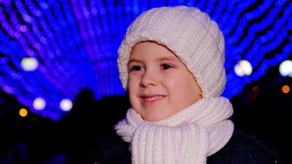 A Handsome Child in a White Hat Smiles Standing Under Christmas Garlands in the Evening