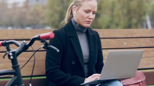 Woman with earphones sitting on bench, using laptop