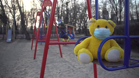 Close-up of Yellow Teddy Bear in Face Mask on Children's Playground with Boy Riding on Swing