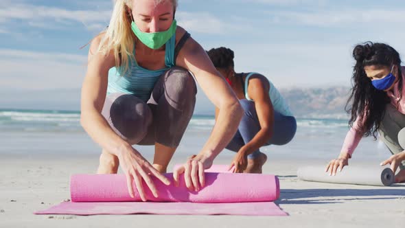 Group of diverse female friends wearing face masks rolling yoga mats at the beach