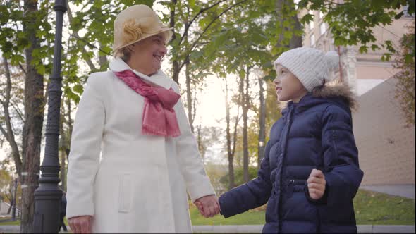 Mature Caucasian Woman in Beige Cloche Hat and White Coat Standing with Her Cheerful Granddaughter