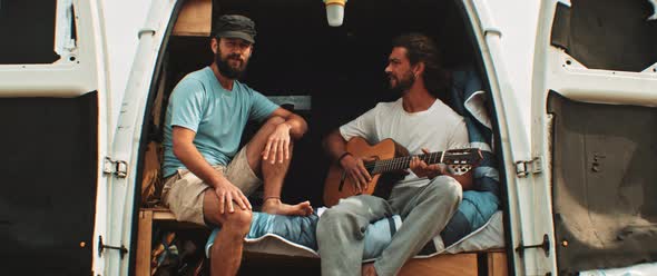 Two friends sitting in back of a van having fun and playing the guitar