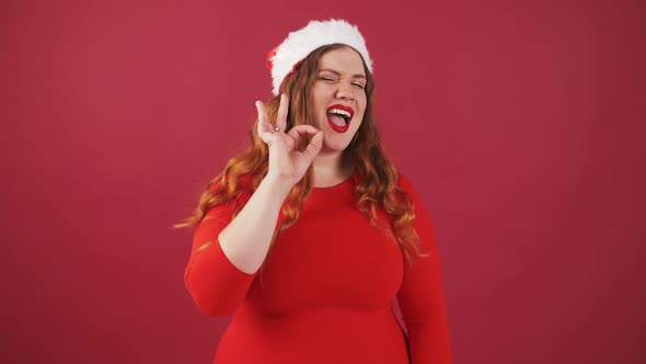 Studio Portrait of Positive Young Oversize Women in Santa Christmas Hat Showing Ok Gesture and