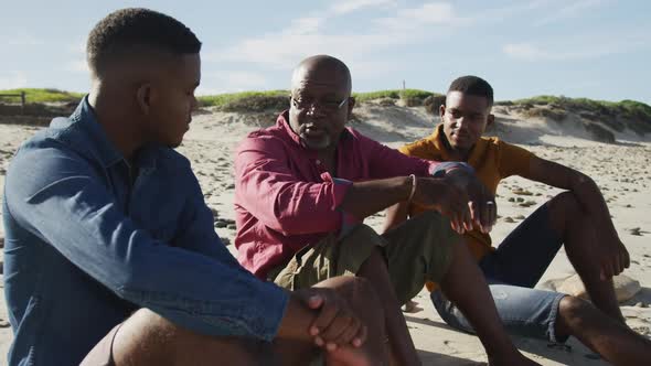African american senior father and twin teenage sons sitting on a beach talking