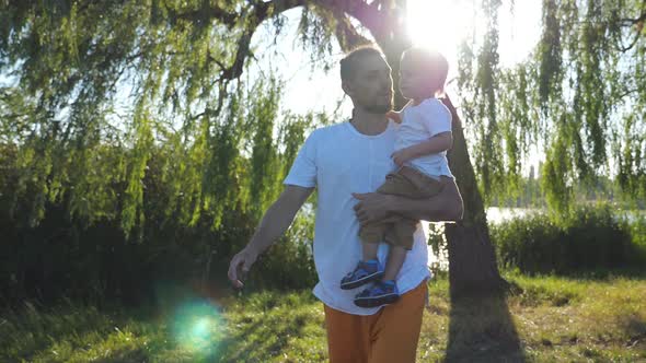 Young Dad Holding His Little Son in Hands and Walking at Summer Park on a Sunny Day