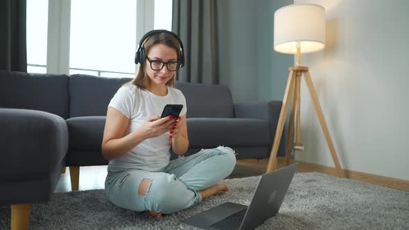 Casually Dressed Woman with Headphones is Sitting on Carpet with Smartphone and Laptop and Working