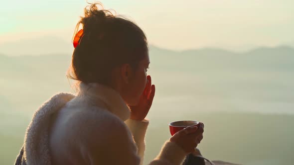Young Woman is Sitting with a Mug of Coffee on a Cliff Overlooking the Mountains with Fog