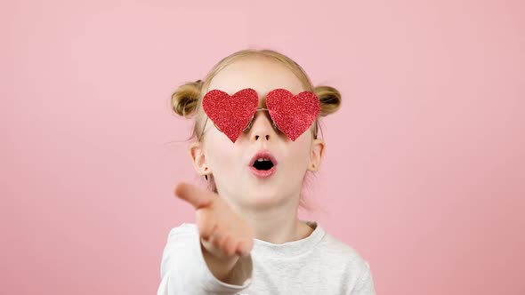 Funny Little Blonde Girl Sending Air Kiss in Red Heart Shape Sunglasses on Pink Background
