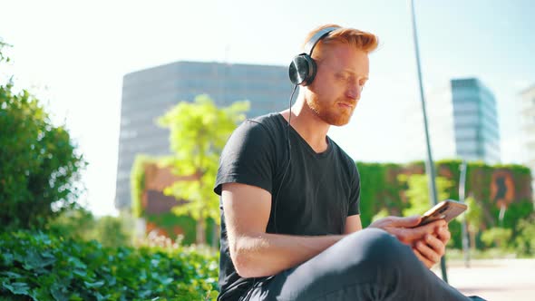 Confident red haired man listening music in headphones and typing by phone