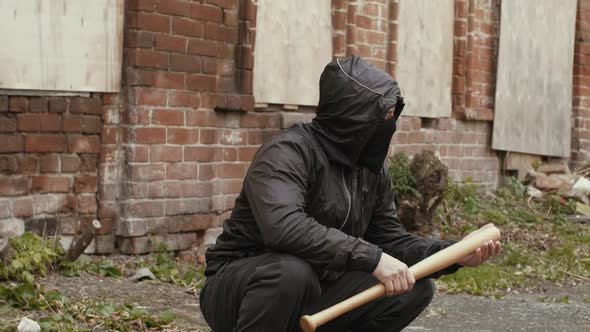 Bandit Man in Black Mask and Jacket with Hood with Baseball Bat Sits on Street