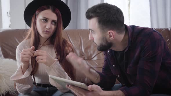 Close-up of Young Bearded Caucasian Man with Tablet Talking To Dissatisfied Pretty Woman in Hat