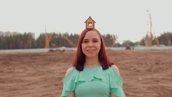 Young Woman with Small Wooden House on Head Stands on Construction Site