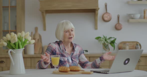 Senior Woman Talking Online By Laptop Sitting at Wooden Kitchen. Smiling Elderly Woman Having Online