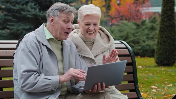 Elderly Married Couple Sit on Bench in Autumn Park Make Video Call Using Laptop Waving Hands Talking