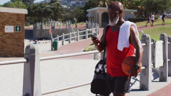 Senior african american man with basketball talking on smartphone while walking near the beach