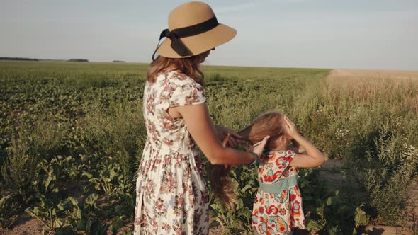 A Mom in a Summer Dress and a Hat Does Her Daughter's Hair By Tying a Ponytail on Her Head While on