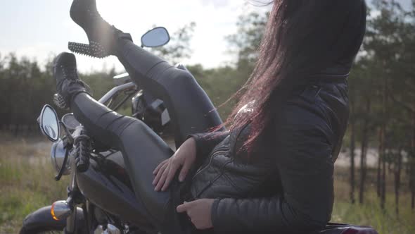 Pretty Young Caucasian Woman in a Black Leather Jacket and Pants Lying on a Classic Motorcycle