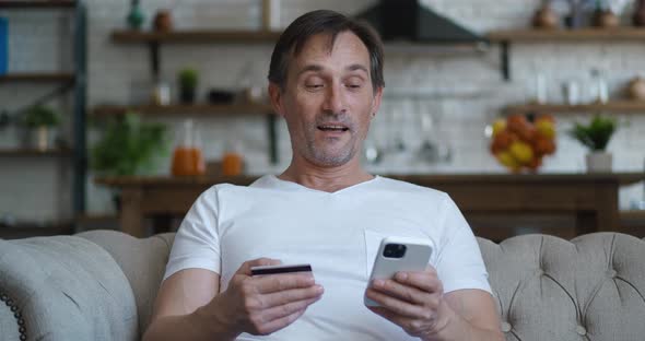 Smiling Mature Man Sitting on Sofa at Living Room Holding Credit Card and Mobile Phone to Make