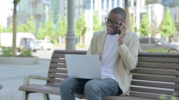 Angry African Man Talking on Phone and Using Laptop While Sitting Outdoor on Bench