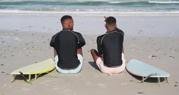 African american teenage twin brothers sitting by surfboards on a beach talking