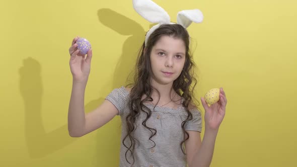 Little Girl in Easter Bunny Ears Holding Two Painted Easter Eggs on a Yellow