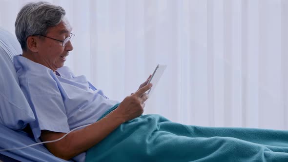 Happy elderly patient sitting on bed and making video call with tablet at hospital