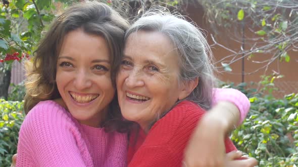 Senior Mother with Gray Hair with Her Adult Daughter Looking at the Camera in the Garden and Hugging