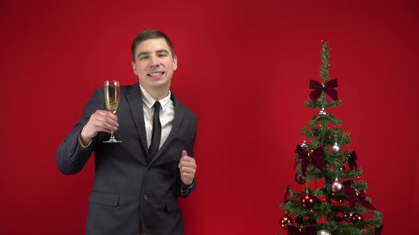 Young Man Dancing with a Glass of Champagne Near the Christmas Tree on a Red Background. A Man in a