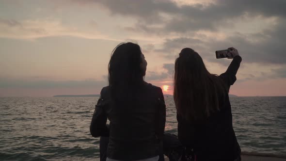 Two girls are taking selfie of themselves near the sea at the sunset