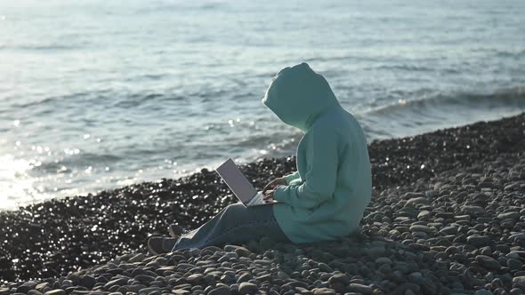 Caucasian Woman Typing on a Laptop on a Rocky Seashore