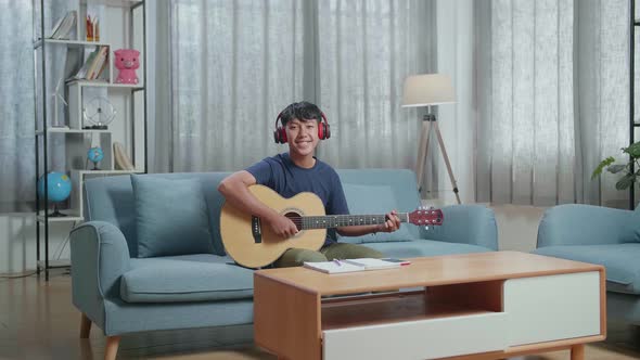 Boy Composer With Notebook On Table Wearing Headphones And Smiling To Camera While Playing Guitar