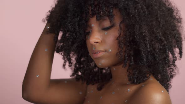 Mixed Race Black Woman with Curly Hair Covered By Crystal Makeup on Pink Background in Studio