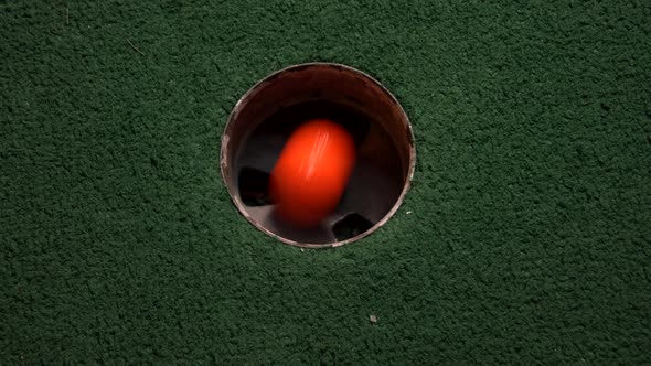 A close up birds eye view of an orange mini golf ball falls into the golf hole and bounces in the po
