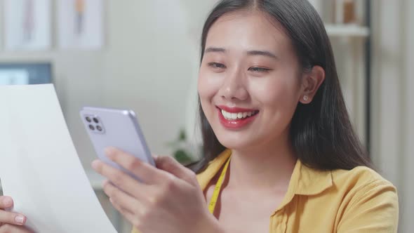 Asian Female Designer Looking At The Picture In Hand And Comparing It To The Pictures On Smartphone