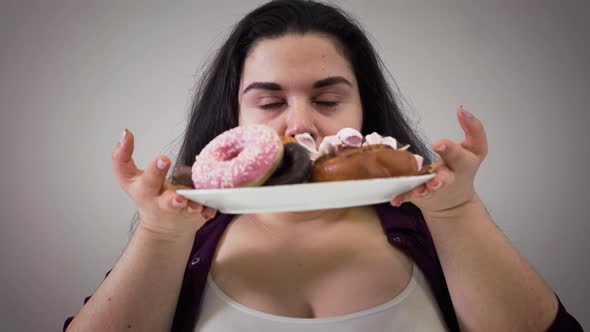Portrait of Hungry Caucasian Fat Woman Taking Plate with Sweets and Smelling Dessert. Obese Girl