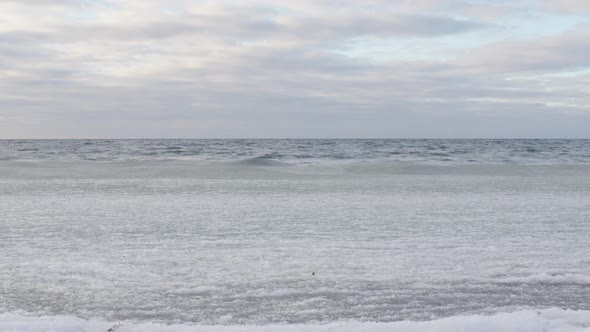 Static shot of ice covered sea waves rolling in on beach. Cloudy blue sky and white palette